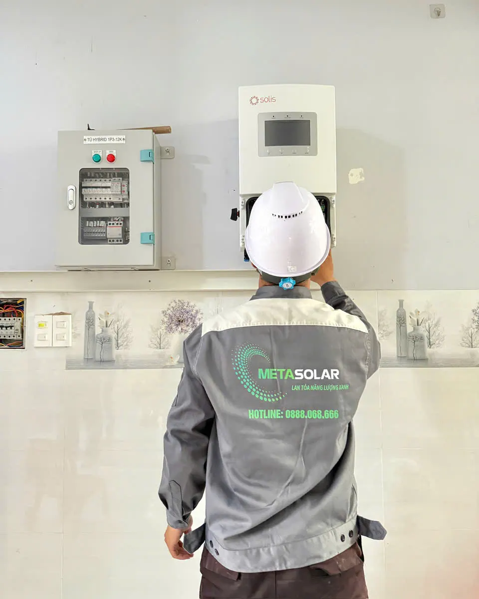 Kỹ thuật viên đội mũ bảo hộ quay lưng thao tác ở đáy inverter Solis cạnh tủ điện hybrid gắn tường